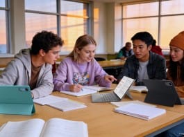 Four students collaborate at a sunlit classroom table with tablets and notebooks across the scene.