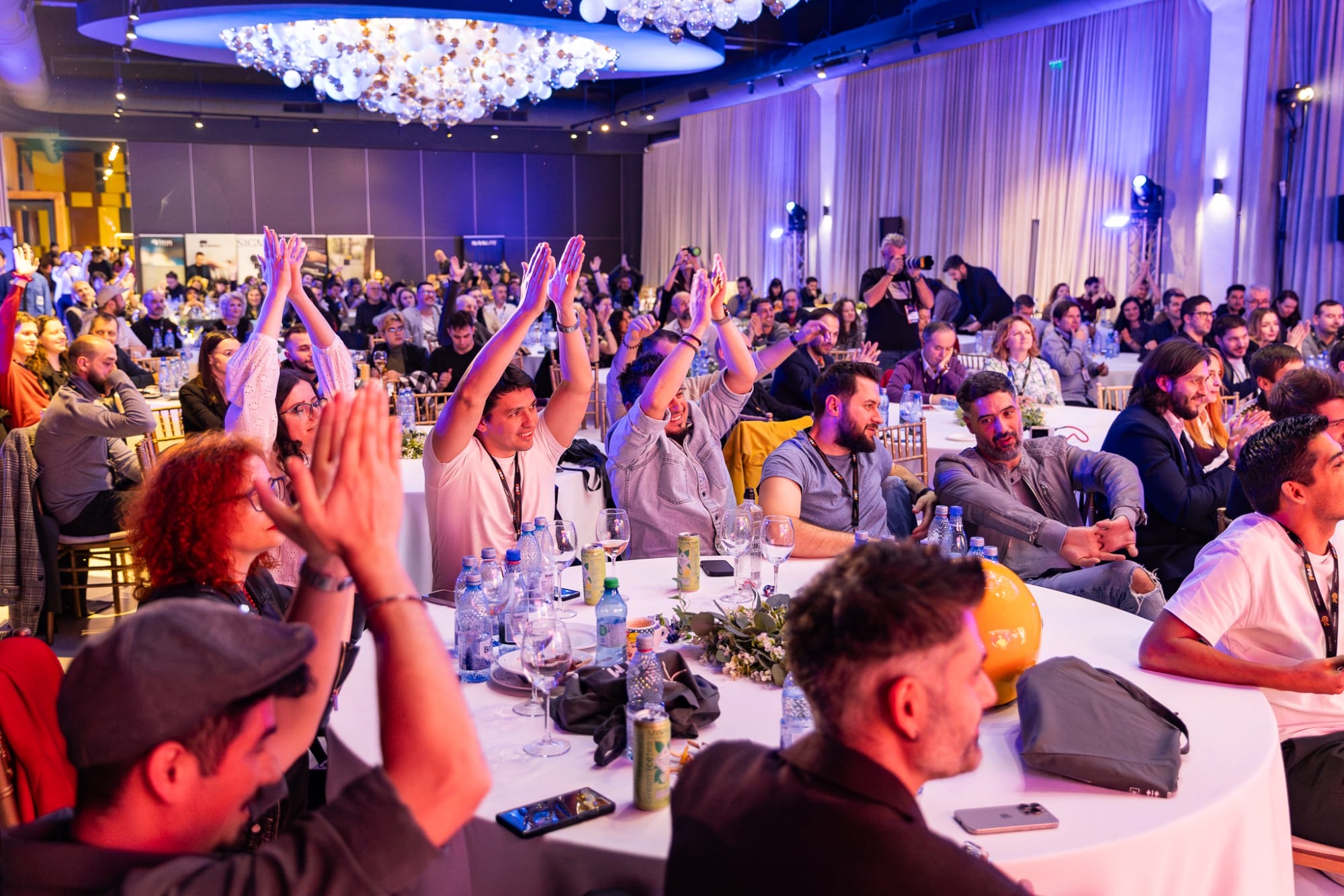 Audience at a formal event clapping around round tables with bottles and decorations, blue lighting overhead.