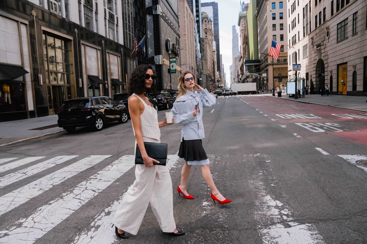 Two stylish women cross a wide city street at a crosswalk: one in a cream jumpsuit holding a black clutch, the other in a striped shirt with a newspaper, red heels, and sunglasses.
