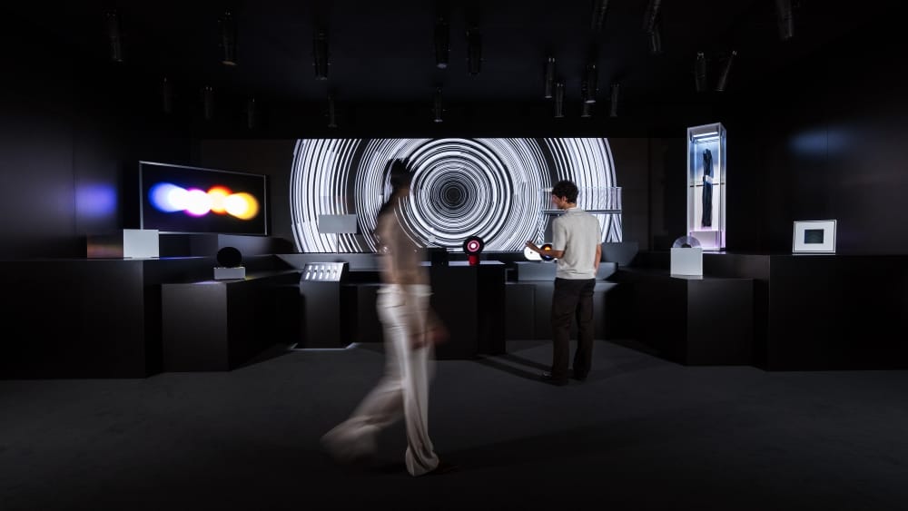 Dim exhibition room with two visitors viewing futuristic displays, including a large circular spiral light installation at the back.