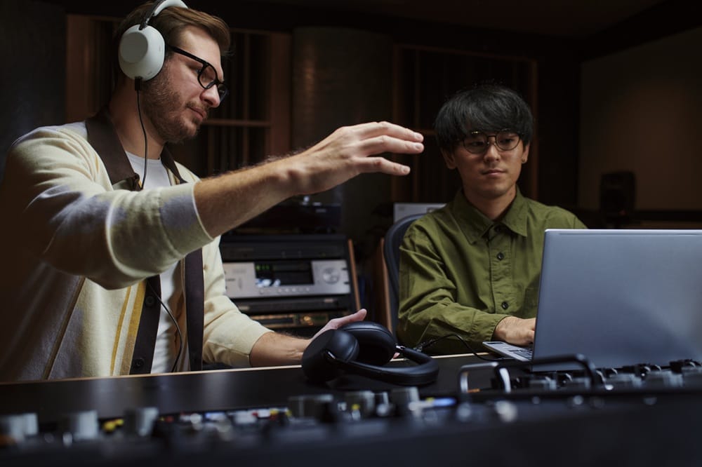 Two men in a recording studio collaborate over a laptop and mixing console; one gestures with his hand while wearing over‑ear headphones.