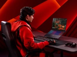 Gamer in a red jacket sits at a desk, playing a PC game on a rainbow-lit laptop with a keyboard and controller nearby, red studio backdrop behind him.
